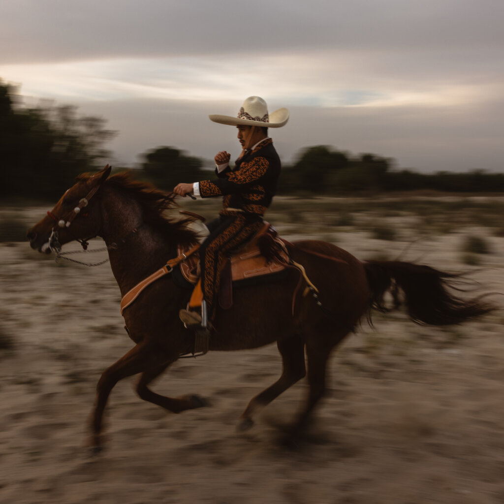A Man In Traditional Mexican Clothing Rides A Brown Horse In A Field