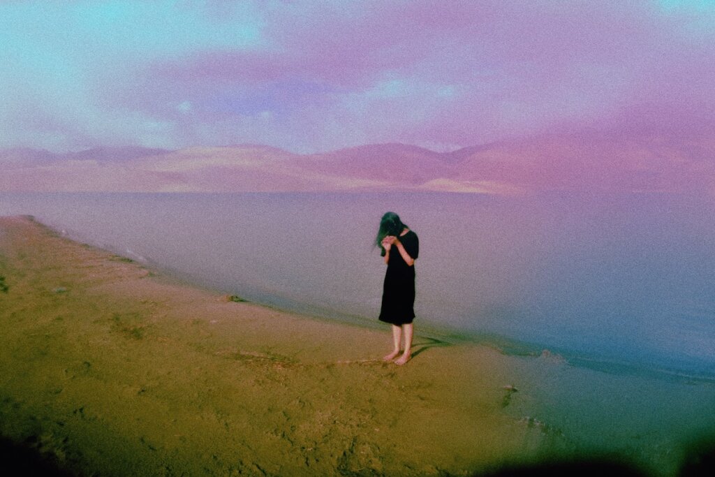 A Teenage Girl Wearing A Black Dress Is Standing Barefoot On The Beach With Mountains In The Distance
