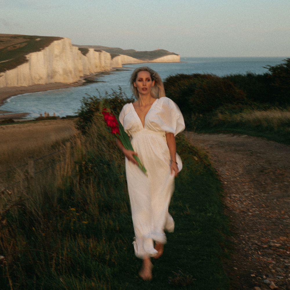 A Woman Is Walking In The Grass Near A Beach with red flowers in hand