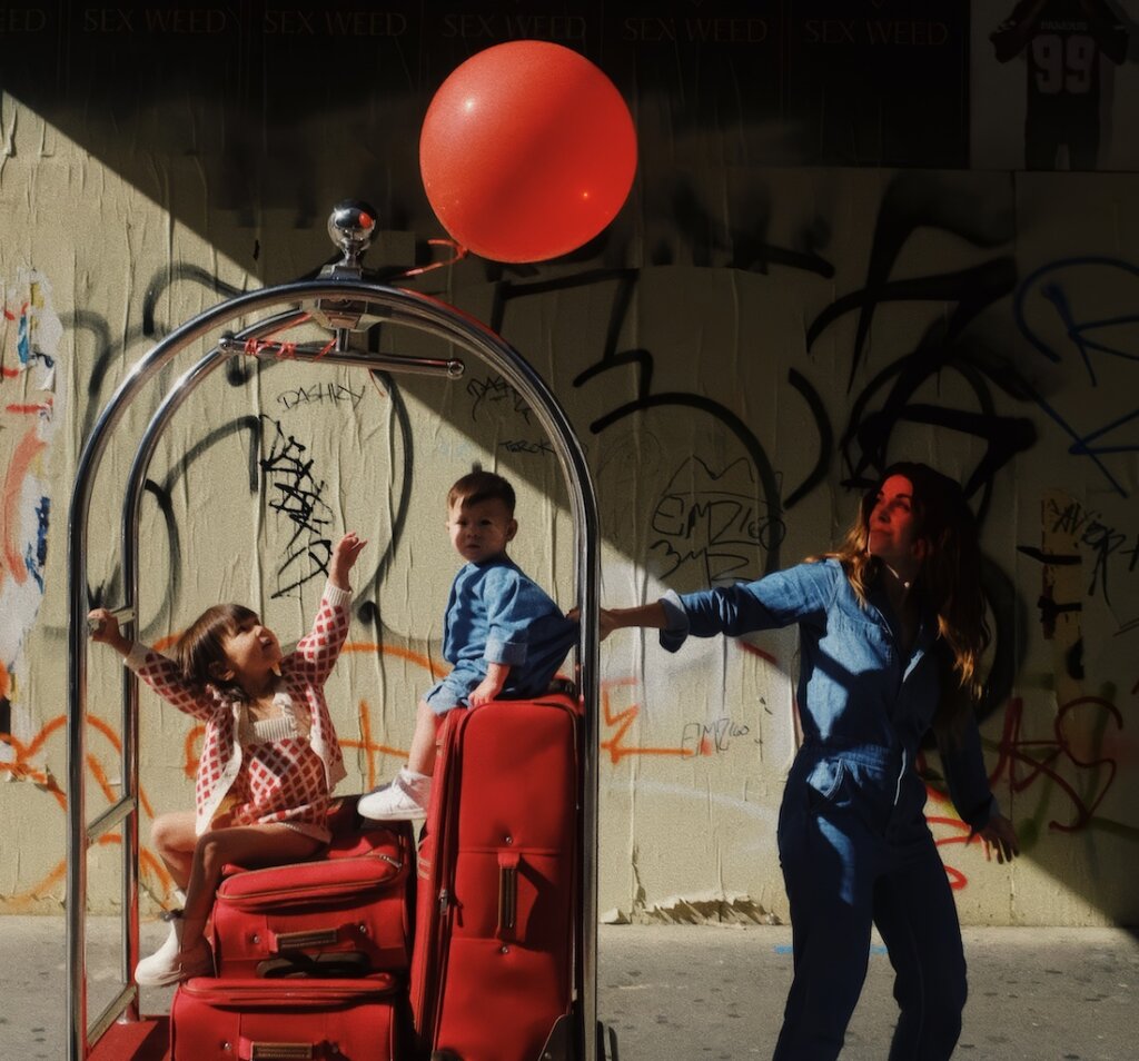 Mother In Denim Clothes Pulling A Cart With Red Suitcases And Her Two Children Sitting With A Red Balloon Tied In The Environment Of A Viaduct Through The City With Graffiti On The Walls