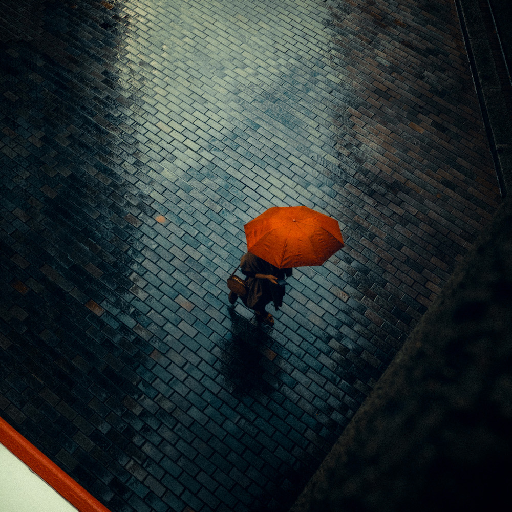 A Person Walks On A Brick Road Holding An Orange Umbrella Under The Rain