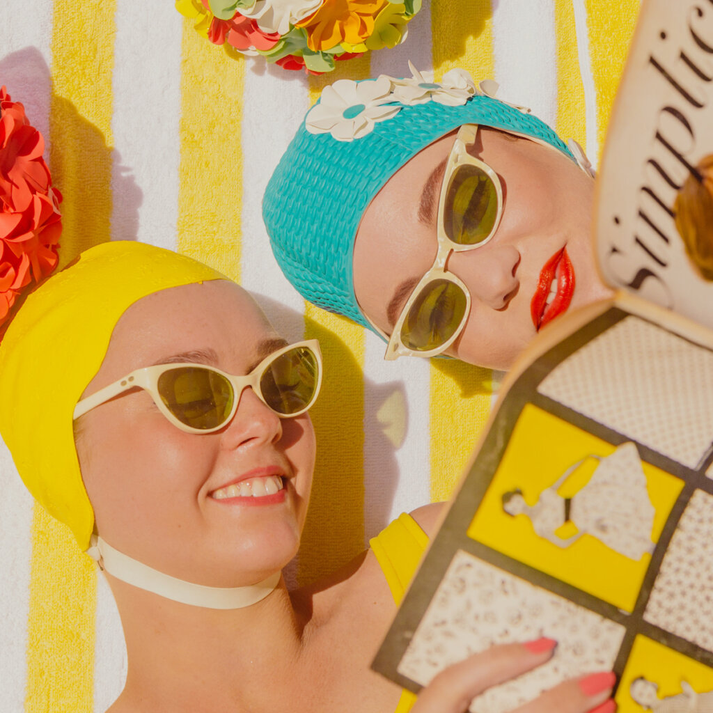 Two Female Friends Smiling With Glasses On, While Laying On Their Backs