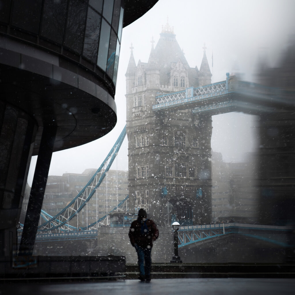 A Man Wearing A Black Hoodie Is Walking In Front Of The Tower Bridge, On A Snowy, Foggy Day