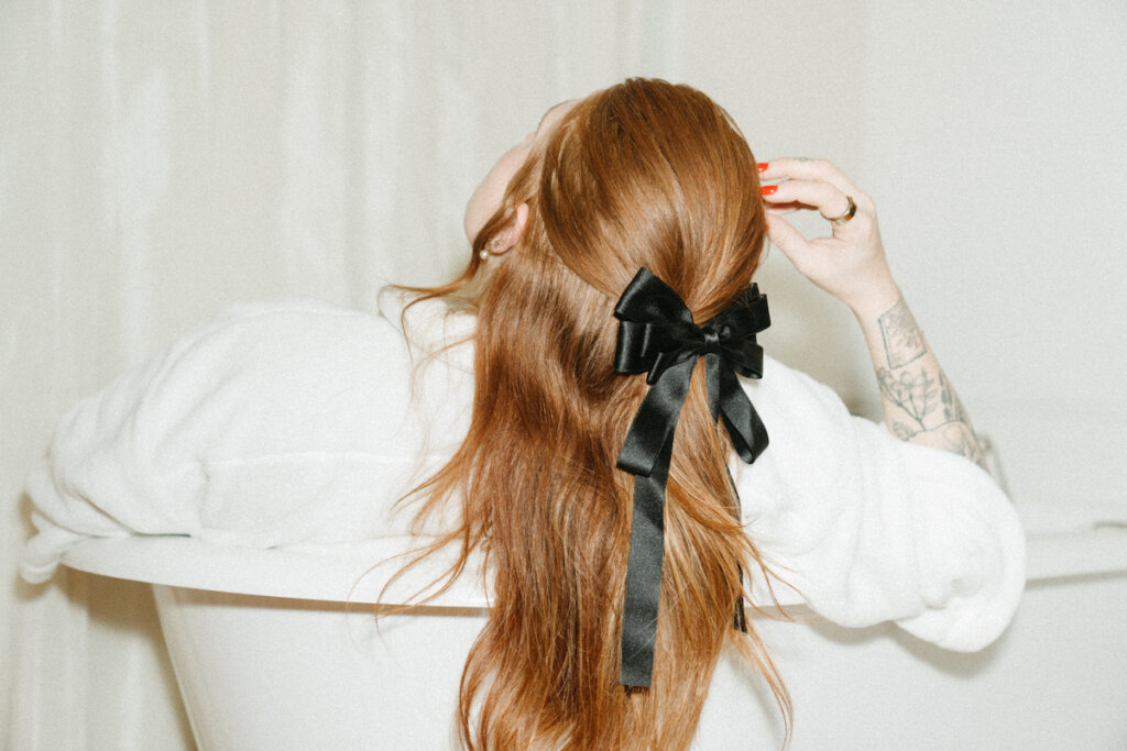 A Woman Wearing White With A Black Bow In Her Long Red Hair Looks Up With Her Hand On Her Head